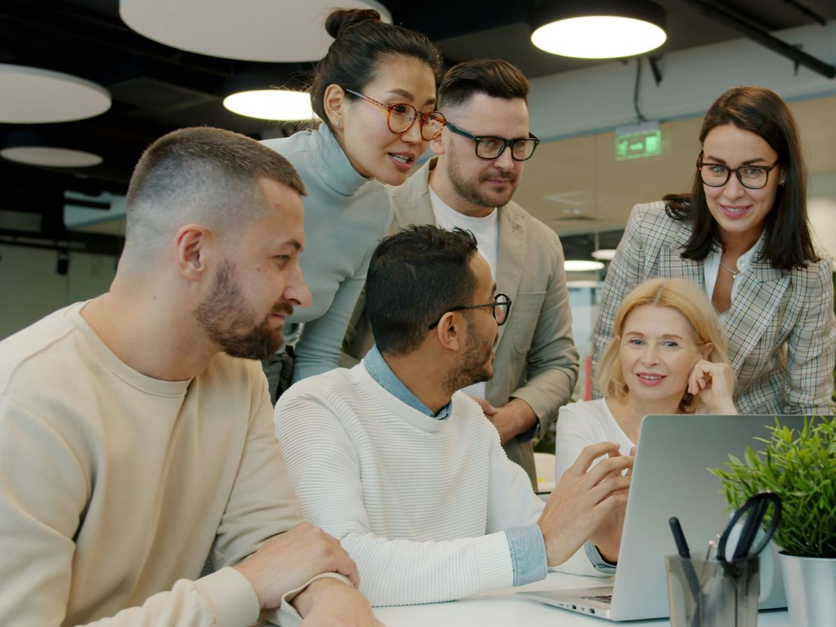 A marketing team of six gathered around one laptop in an open-plan office