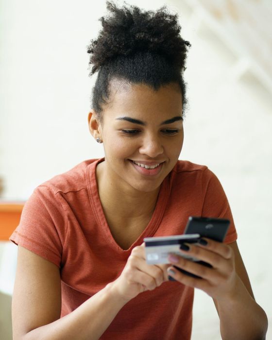 A smiling young woman holding a bank card and a smartphone at home