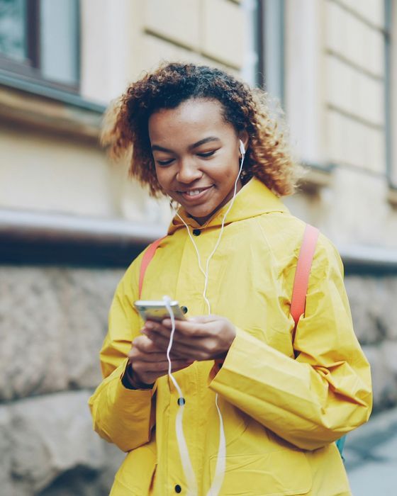 A young woman in a yellow raincoat using her smartphone with earphones on the street