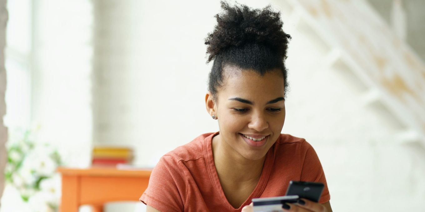 A smiling young woman holding a bank card and a smartphone at home