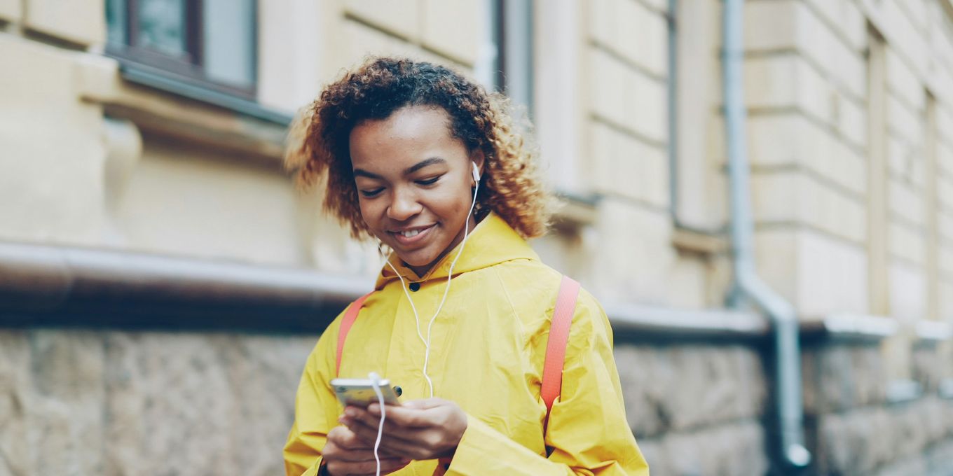 A young woman in a yellow raincoat using her smartphone with earphones on the street
