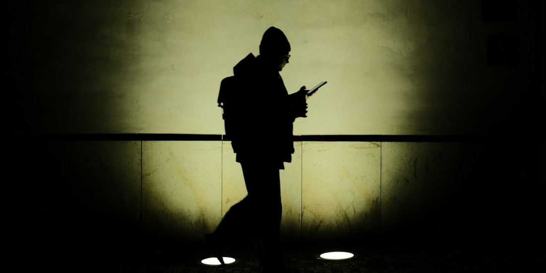 Silhouette of a person walking and looking at a phone against a lit wall at night